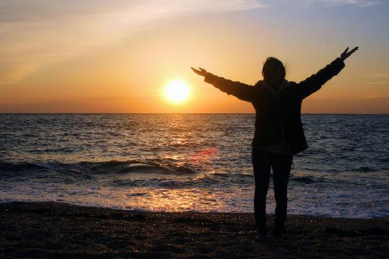 Photograph of Autumn standing on the shore facing the ocean with her arms outstretched, silhouetted against a vibrant sunset. The sun is low on the horizon, casting a warm orange and yellow glow across the sky and reflecting on the water. Waves are gently rolling onto the beach, and the overall scene conveys a sense of peace and freedom.